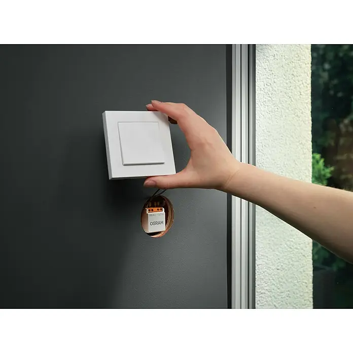 Osram Smart+ Schalter 1CH - Weiß, Innen - Eine Hand montiert einen weißen OSRAM-Lichtschalter an einer grauen Wand. Der Schalter ist quadratisch und hat einen kleineren quadratischen Knopf.