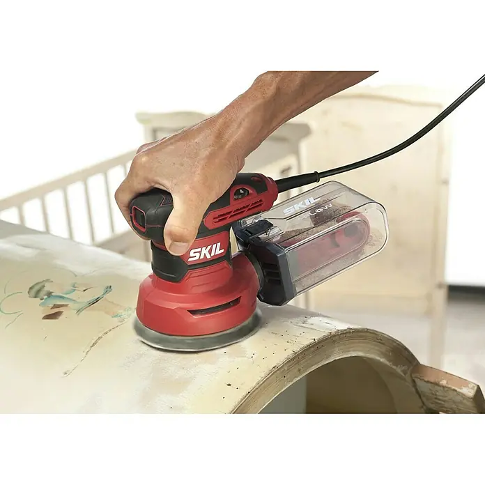 Skil Elektro-Exzenterschleifer 7471 AA - 300 W, Durchmesser Schleifteller: 125 mm - Nahaufnahme eines SKIL Exzenterschleifers mit Staubbehälter, der auf einer beigen Oberfläche verwendet wird.
