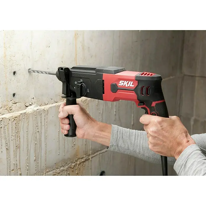 Skil Kombihammer 1770 GA - 850 W, 2,8 J, SDS-Plus-Aufnahme - Nahaufnahme einer roten SKIL Schlagbohrmaschine, die in eine Betonwand bohrt, mit Bohrlöchern sichtbar.
