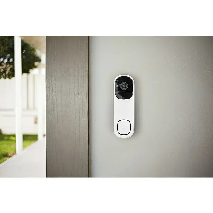 Osram Smart+ Türklingel mit Kamera - Weiß - Eine Nahaufnahme einer smarten, weißen Türklingelkamera, die an einer grauen Wand neben einem Türrahmen montiert ist.