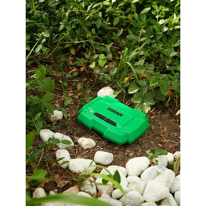 Osram Smart+ Bodenfeuchtesensor - Grün - Grüne OSRAM-Gerätebox liegt auf dem Boden zwischen Steinen und Pflanzen. Ideal für Outdoor-Aktivitäten und sichere Aufbewahrung.