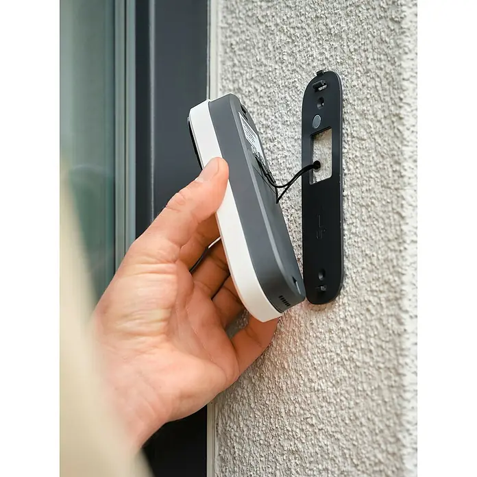 Osram Smart+ Türklingel mit Kamera - Weiß - Eine smarte Türklingel wird an einer Montagehalterung an einer Außenwand befestigt. Die Klingel ist grau mit weißen Akzenten.