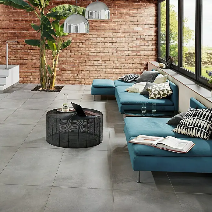 Art-Tec by Palazzo Sockelfliese - 7 x 60 cm, Steel, Matt - Modernes Wohnzimmer mit blauem Ecksofa, rundem Gittertisch, Steinfliesen und Backsteinwand für ein gemütliches Ambiente.