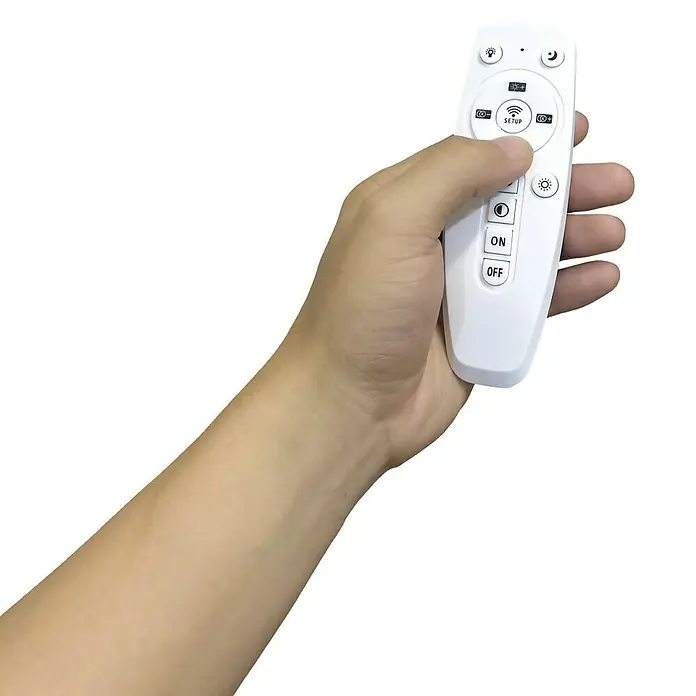 Panaled Plafón LED Katto II - 110 W, L x An x Al: 89,5 x 29,5 x 4 cm, Blanco, Blanco cálido - Una mano sujeta un mando a distancia blanco. El dispositivo tiene botones para encender y apagar, y ajustar el brillo.