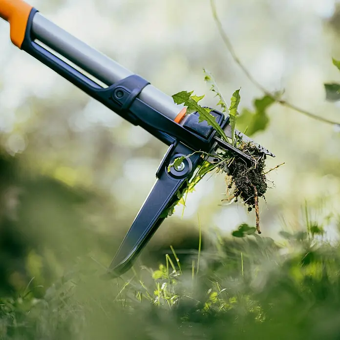 Fiskars X-series Unkrautstecher - Länge: 1.100,7 mm - Nahaufnahme eines Unkrautstechers mit langem Griff, der Unkraut aus einem Rasen entfernt. Das Werkzeug hat einen orangefarbenen Griff und einen grauen Schaft.
