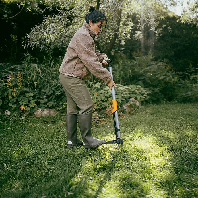 Fiskars Unkrautstecher - Länge: 106,2 cm - Eine Frau benutzt einen manuellen Unkrautstecher mit langem Griff im Garten, um Unkraut zu entfernen. Das Werkzeug ist grau mit orangefarbenen Akzenten.