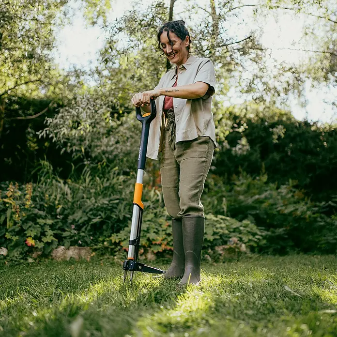 Fiskars X-series Unkrautstecher - Länge: 1.100,7 mm - Eine Frau benutzt einen Fiskars Unkrautstecher im Garten. Das Werkzeug hat einen schwarzen Griff und einen orangefarbenen Akzent.