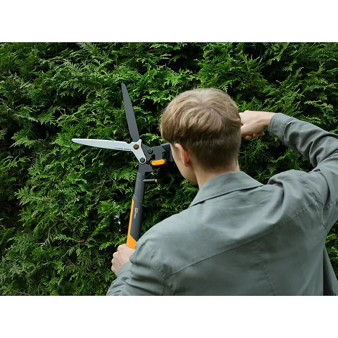 Fiskars X-series Hand-Heckenschere DualAction HSX96 - Länge: 62,7 cm, orange, schwarz, metallic - Eine Person schneidet eine Hecke mit einer Fiskars Heckenschere. Die Schere hat lange Griffe für mehr Reichweite und Hebelwirkung.