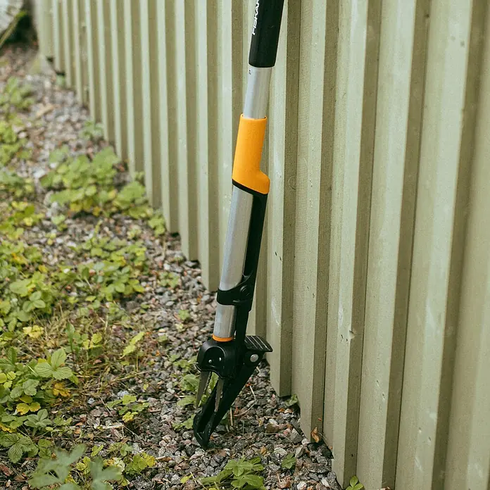 Fiskars Unkrautstecher - Länge: 106,2 cm - Das Bild zeigt einen Fiskars Unkrautstecher, ein Werkzeug zum einfachen Entfernen von Unkraut, das an einem Zaun lehnt und von Unkraut umgeben ist.