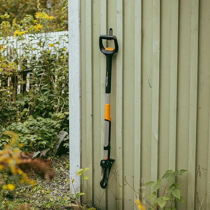 Fiskars X-series Unkrautstecher - Länge: 1.100,7 mm - Eine Fiskars Unkrautentfernungswerkzeug hängt an einer hölzernen Wand, mit einem schwarzen Griff und orangefarbenen Akzenten, ideal für die Gartenarbeit.