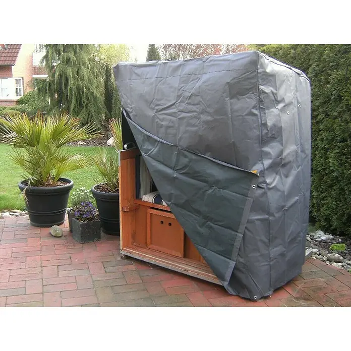 Westerholt Strandkorb-Schutzhülle - L x B x H: 135 x 100 x 134 cm, 420D Oxford Polyester, Anthrazit - Nahaufnahme einer grauen Schutzhülle für einen Strandkorb auf einer Terrasse, umgeben von Pflanzen und einem Haus im Hintergrund.