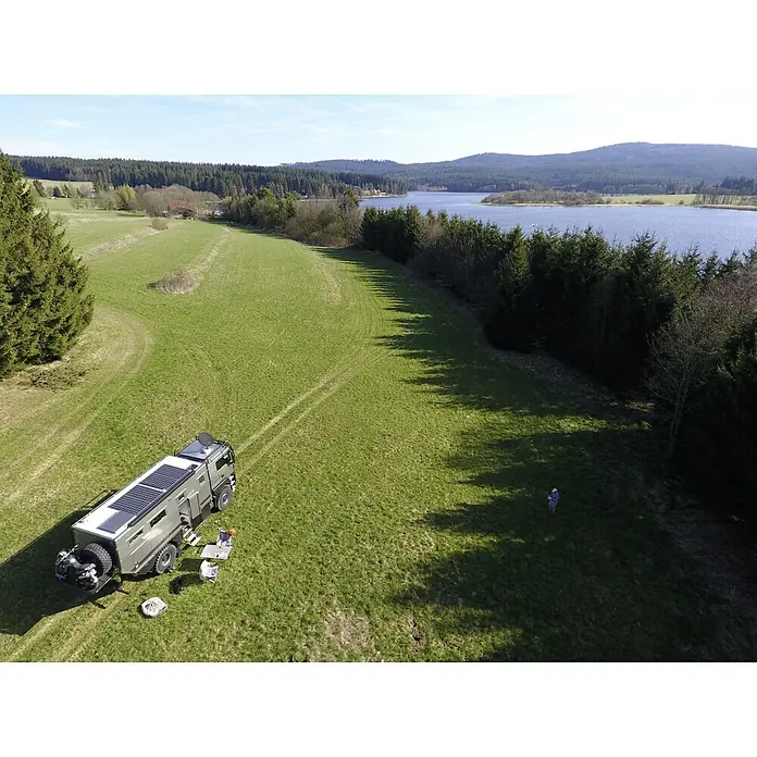 Sunset Solarmodul-Set Caravan - Nennleistung: 140 W, Für den Caravan- und Campingbereich - Ein Expeditionsmobil mit Solaranlage auf einem grünen Feld mit Blick auf einen See. Ein paar Stühle und ein Tisch stehen draußen.