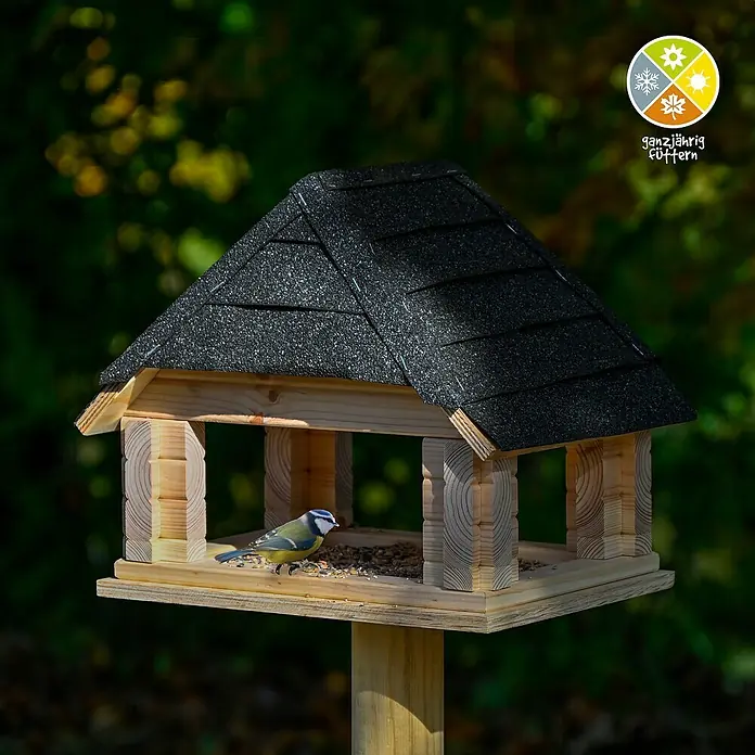 Dobar Vogelhaus Sort - Holz, Natur/Schwarz - Vogelhaus mit schwarzem Dach und kleinem Vogel, der auf dem Futterbrett sitzt. Das Vogelhaus ist aus hellem Holz gefertigt und steht auf einem Pfosten.