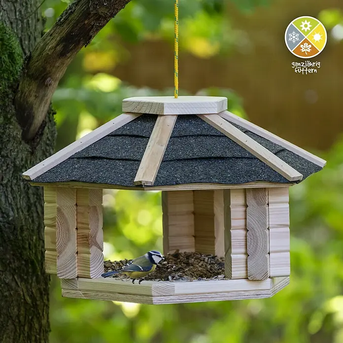 Dobar Vogelhaus Zwart - Holz, Natur/Schwarz - Ein hölzernes Vogelhaus mit einem kleinen Vogel darin, das an einem Baum hängt, mit einem "ganzjährig füttern" Logo in der Ecke.
