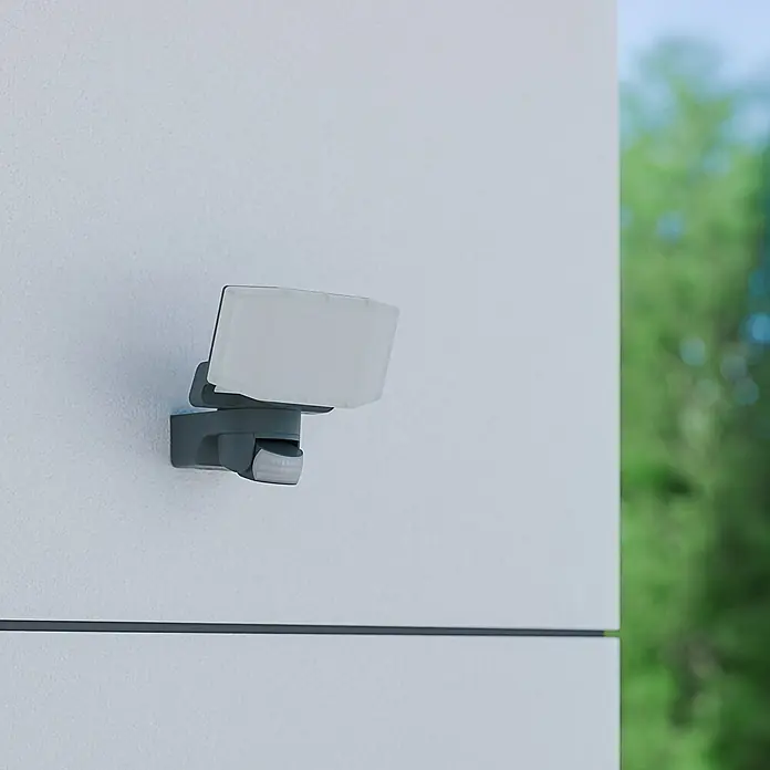 Steinel LED-Sensor-Außenwandleuchte XLED Protect S - 14,3 W, Anthrazit, Warmweiß - Diese Nahaufnahme zeigt einen grauen, nach außen gerichteten LED-Strahler mit Bewegungsmelder, der an einer weißen Wand montiert ist.