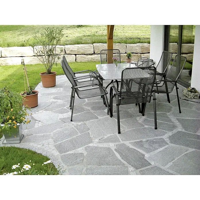 Polygonalplatte - Quarzit, Lieferumfang: ca. 10 m² - Ein einladender Terrassenbereich mit einem eleganten Tisch und Stühlen auf einer stilvollen Steinplattenfläche, umgeben von üppigem Grün.
