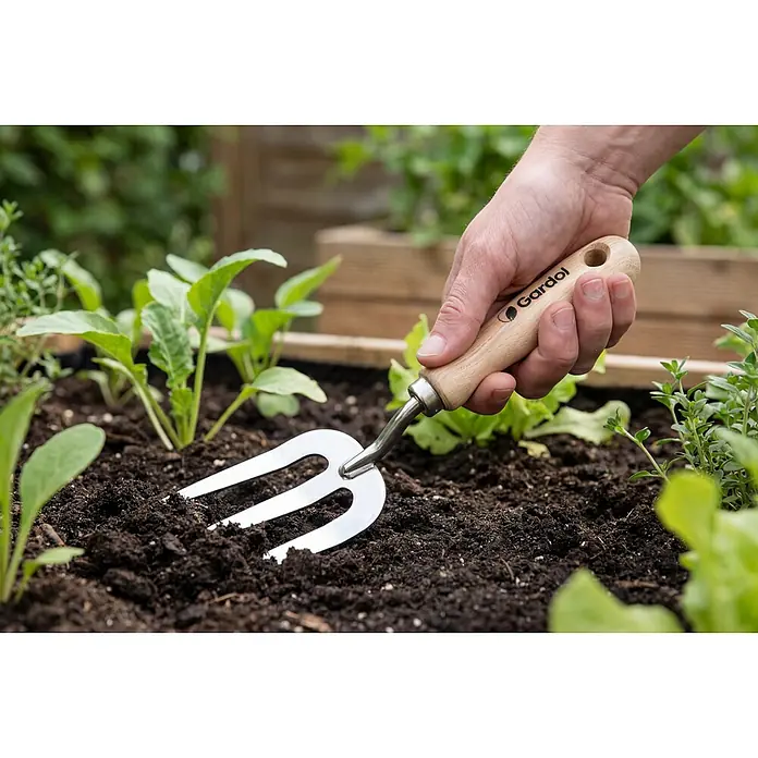 Gardol Spatengabel - Anzahl Zinken: 3 Stk. - Nahaufnahme einer Hand, die eine Gardol Gartenforke in dunkle Gartenerde sticht, umgeben von grünen Pflanzen.