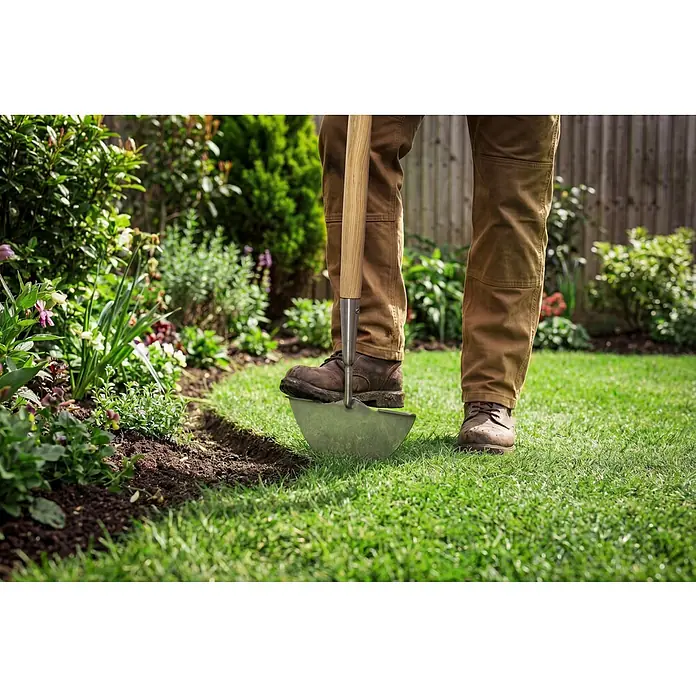 Gardol Rasenkantenstecher - Esche, Edelstahl - Eine Person bearbeitet einen Garten mit einem Spaten, der in den Rasen gesteckt wird. Im Hintergrund sind Pflanzen und ein Zaun sichtbar.