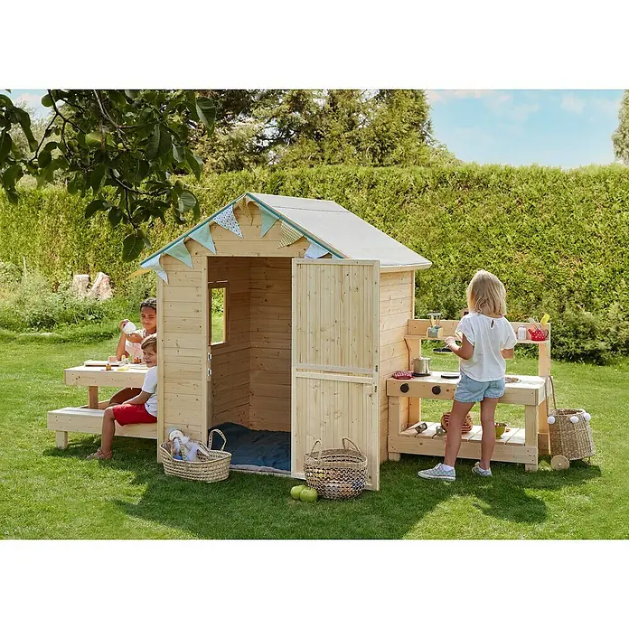 Soulet Spielhaus Blanche - L x B x H: 302 x 127 x 162 cm, Holz - Ein hölzernes Spielhaus mit Küche und Sitzbereich im Garten. Kinder spielen rund um das Spielhaus, das mit bunten Wimpeln dekoriert ist.