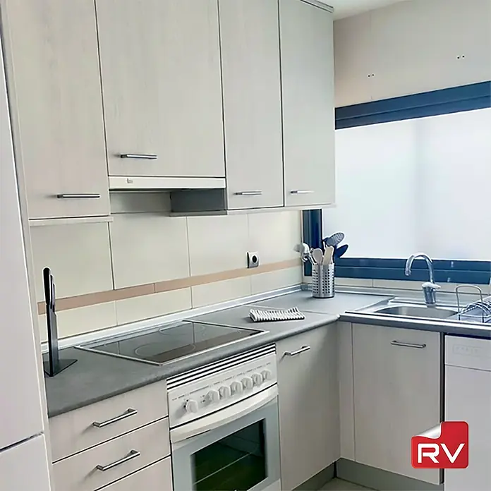 Lámina decorativa autoadhesiva vinílico autoadhesivo a metros - Madera decapada blanca - Una cocina luminosa con armarios blancos, una encimera gris, una cocina y un fregadero, todo ello situado bajo una ventana con marco oscuro.