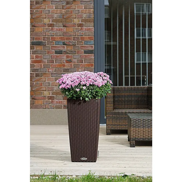 Lechuza Cottage Pflanzsäule eckig Cubico 40 - Außenmaß (L x B x H): 39,5 x 39,5 x 75,5 cm, Kunststoff, Mokka - Ein hoher, brauner Lechuza Pflanzkübel mit üppigen rosa Blüten steht auf einer Terrasse vor einer Ziegelwand und einem Terrassenset.