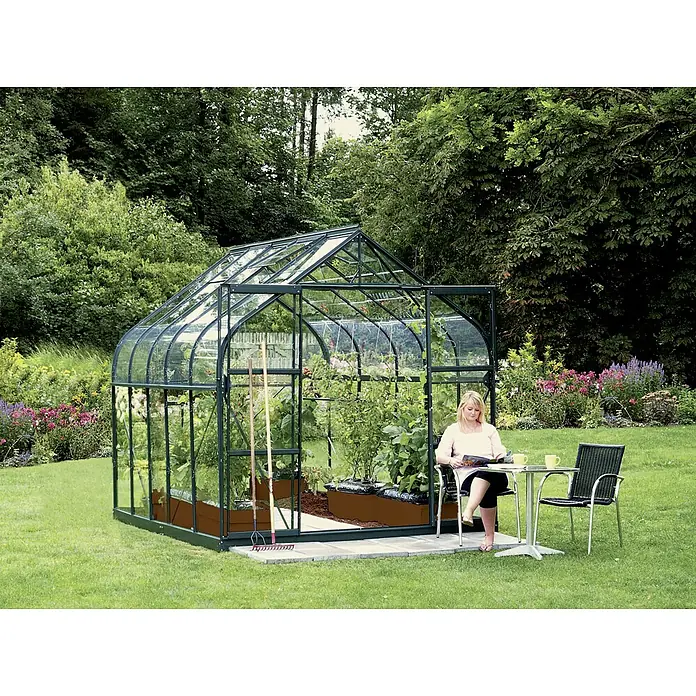 Vitavia Gewächshaus Dione 8300 - Außenmaß inkl. Dachüberstand (B x T): 263,5 x 321 cm, 3 mm, Smaragd - Eine Frau sitzt neben einem Gewächshaus im Garten und liest. Das Gewächshaus ist grün und hat Pflanzen darin.