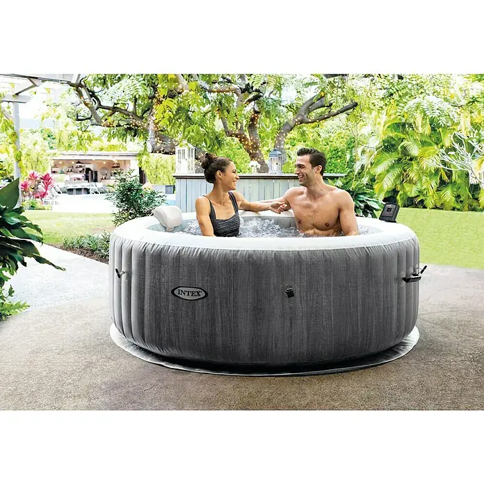 Intex Pure Spa Hidromasaje móvil Greenwood - Ø x Al: 196 x 71 cm, Gris/Blanco, 795 l - Una bañera de hidromasaje Intex inflable instalada en un jardín con una pareja relajándose en ella. La tina gris está en un patio bajo los árboles.