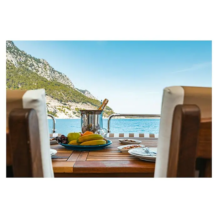 Marine Business Cocoa Figura decorativa Ballena - Azul/Marrón, Madera, 1 ud. - Una mesa de yate con una cubitera, fruta variada y platos, con un paisaje costero de fondo.