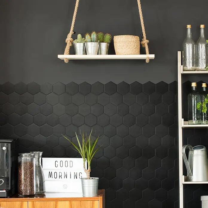 Smart Tiles Selbstklebemosaik Hexa - 24,78 x 23,75 cm, Walton, Matt - Stilvolles Interieur mit einer schwarzen Sechseckfliesenwand, einem hängenden Regal mit Kakteen und einer "Good Morning"-Leuchtreklame.