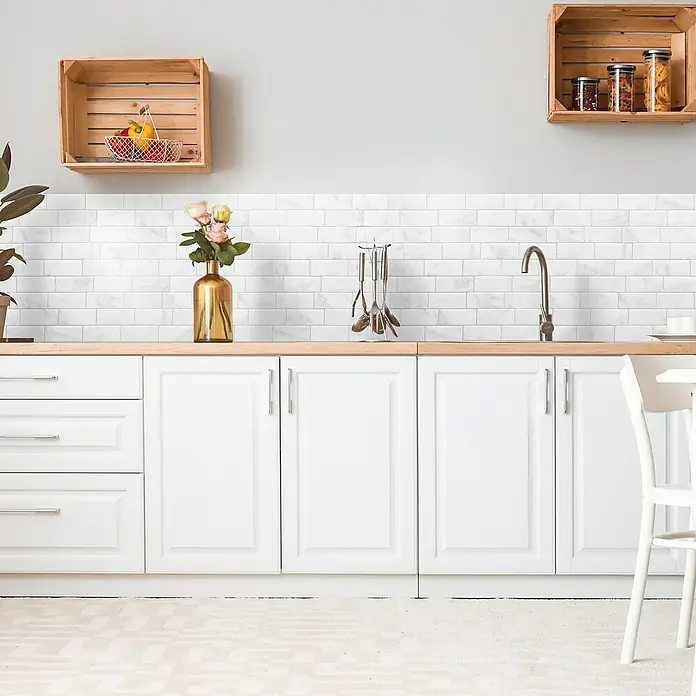 Smart Tiles Selbstklebemosaik Metro - 29,34 x 21,28 cm, Carrera, Glänzend - Küchenansicht mit weißen Schränken, einer Holzarbeitsplatte, weißen Fliesen und Dekorationen wie einem Blumenstrauß und Küchenutensilien.