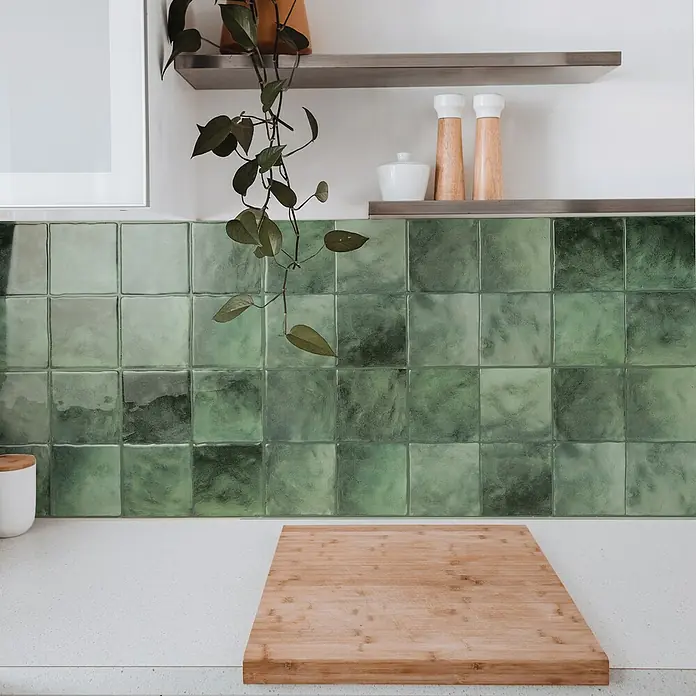 Smart Tiles Selbstklebemosaik Zellige - 22,86 x 22,86 cm, Taza, Glänzend - Eine rechteckige Bambus-Schneidebrett auf einer weißen Küchentheke vor einem grünen Fliesenwandbild, das eine praktische Küchenausstattung zeigt.