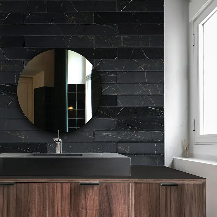 Smart Tiles Selbstklebemosaik Norway - 57,31 x 49,4 cm, Dorato, Matt - Badezimmerwaschtisch mit schwarzem Marmorwaschbecken und runden Spiegel vor einer Wand mit schwarzen Fliesen.