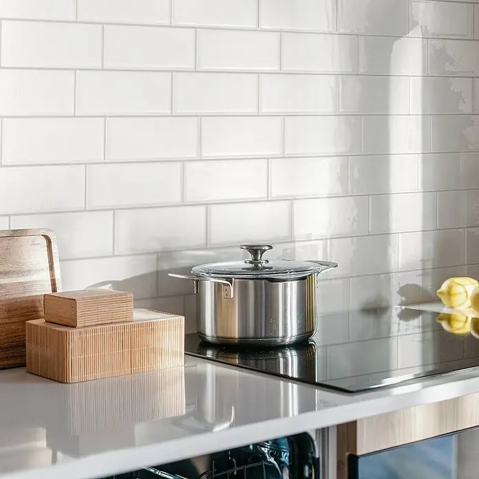 Smart Tiles Selbstklebemosaik XL Metro - 53,47 x 23,05 cm, Chicago, Glänzend - Küchenstillleben mit einem Topf auf einem Kochfeld, umgeben von Holzaccessoires und frischen Zitronen vor einem weißen Fliesenspiegel.