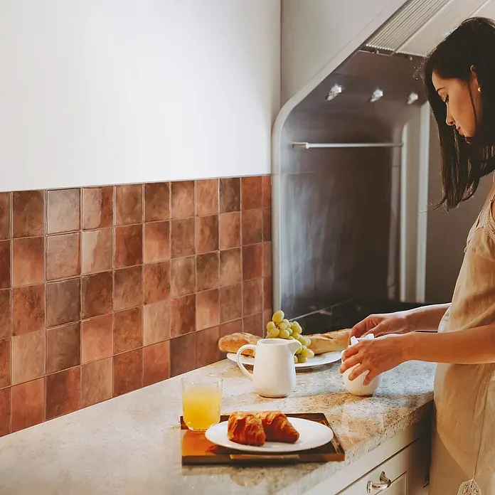 Smart Tiles Selbstklebemosaik Zellige - 22,86 x 22,86 cm, Safi, Glänzend - Eine Frau bereitet Frühstück in einer Küche mit braunen Fliesen und Granitarbeitsplatten zu. Ein Glas Saft, Croissants und Obst sind auf dem Tisch.