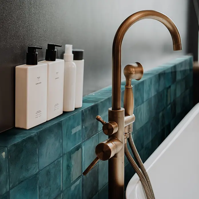 Smart Tiles Selbstklebemosaik Zellige - 22,86 x 22,86 cm, Costa, Glänzend - Elegante Badarmatur aus Bronze mit stilvollen Pflegeprodukten im Hintergrund. Ein luxuriöses Badezimmerdetail.