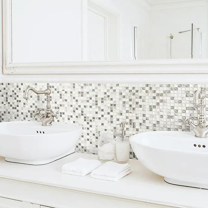 Smart Tiles Selbstklebemosaik Minimo - 29,34 x 24,48 cm, Noche, Glänzend - Ein Badezimmerwaschtisch mit zwei Aufsatzwaschbecken, Armaturen und einem Spiegel mit Mosaikfliesen-Akzentwand.