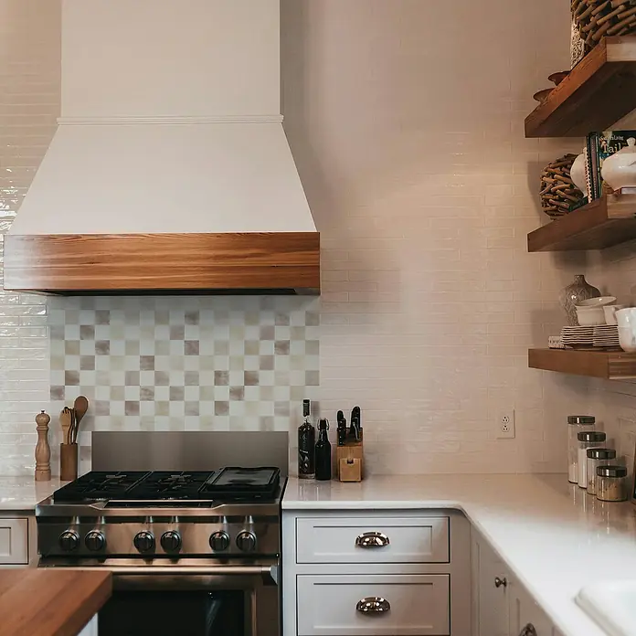Smart Tiles Selbstklebemosaik Damier - 19,69 x 19,69 cm, Luxor, Matt - Einbauküche mit Edelstahlherd, Dunstabzugshaube mit Holzrand und hellen Fliesen. Regale präsentieren Geschirr und Gewürze.