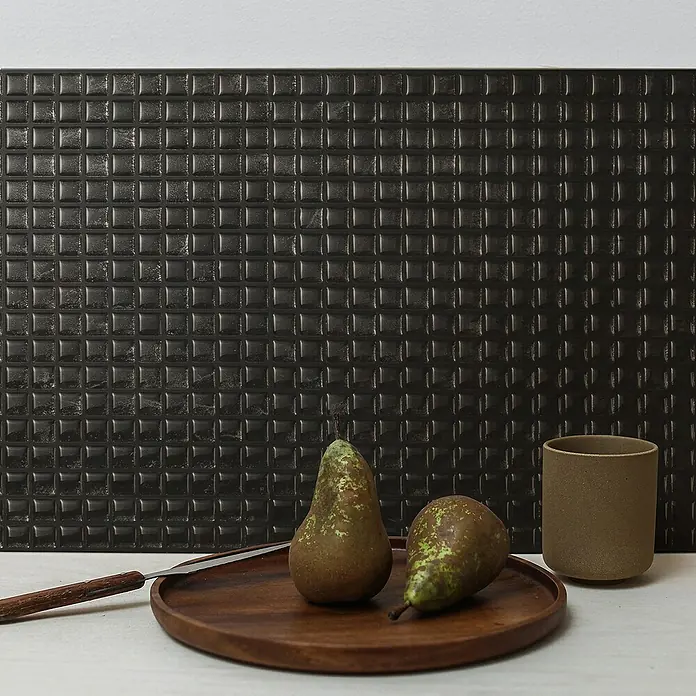 Smart Tiles Selbstklebemosaik Minimo - 29,34 x 24,48 cm, Nero, Glänzend - Stillleben mit zwei Birnen, einem Messer und einer Tasse auf einem Holzteller vor einer dunklen Mosaikwand.
