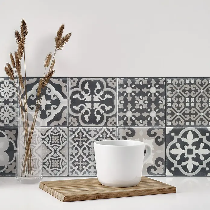 Smart Tiles Selbstklebemosaik Vintage - 22,86 x 22,86 cm, Arezzo, Glänzend - Nahaufnahme einer weißen Tasse auf einem Holzbrett vor gemusterten Fliesen, ergänzt durch dekoratives Gras in einer Glasvase.