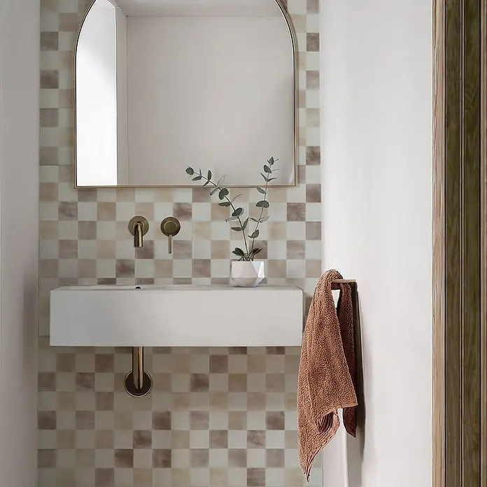 Smart Tiles Selbstklebemosaik Damier - 19,69 x 19,69 cm, Luxor, Matt - Badezimmer mit quadratischen Fliesen. Ein weißes Waschbecken und ein goldener Spiegel mit abgerundeten Ecken sind dekorative Elemente.