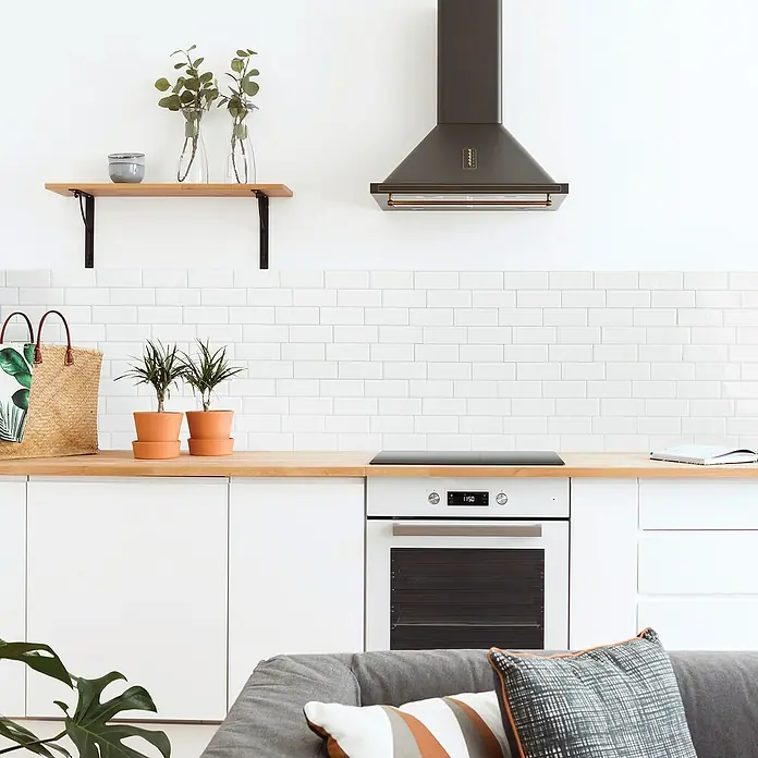 Smart Tiles Selbstklebemosaik Metro - 29,34 x 21,28 cm, Campagnola, Glänzend - Küchenszene mit Dunstabzugshaube, weißen Schränken, Holzregal, Zimmerpflanzen, Tasche und Couch mit Kissen in einem hellen Raum.