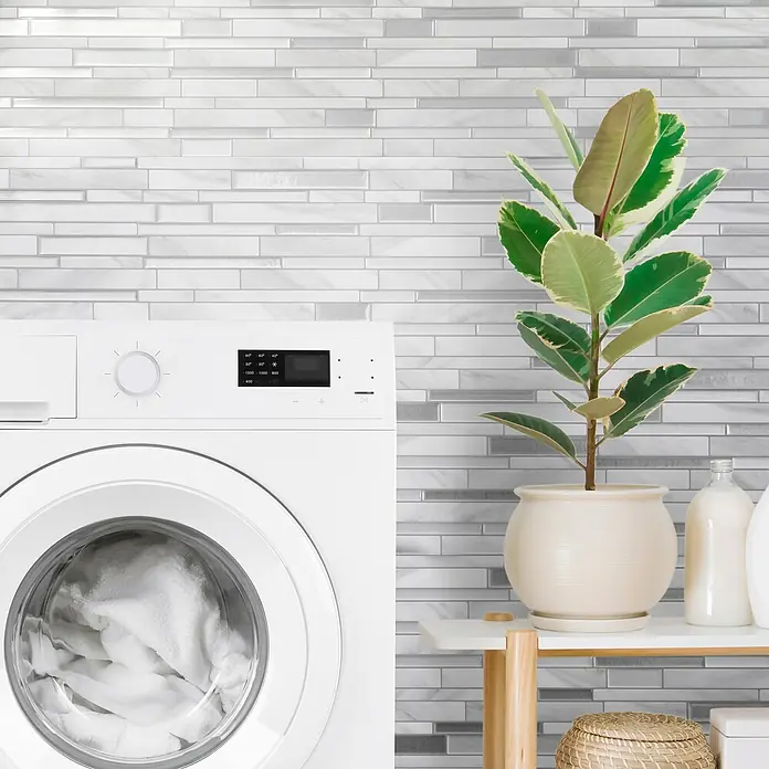 Smart Tiles Selbstklebemosaik Milano - 29,33 x 24,45 cm, Carrera, Matt - Nahaufnahme einer weißen Waschmaschine mit silberfarbenen horizontalen Wandfliesen im Hintergrund, die ein modernes und sauberes Erscheinungsbild vermitteln.