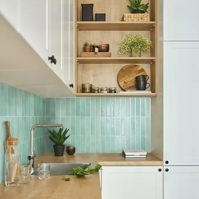 Smart Tiles Selbstklebemosaik Morocco - 29,02 x 22,86 cm, Asilah, Glänzend - Einblick in eine moderne Küche mit mintgrünen Fliesen, hellen Holzflächen und weißen Schränken. Dekorative Elemente wie Pflanzen und Geschirr ergänzen das Design.