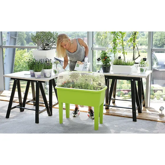 Hochbeet Respana Planter Set - Steingrau, 77 x 58 x 96,3 cm, Kunststoff - Frau, die einen grünen erhöhten Pflanzkasten in einem Gewächshaus pflegt, mit Pflanzen, Töpfen und Tischen für eine Indoor-Gartenarbeit.