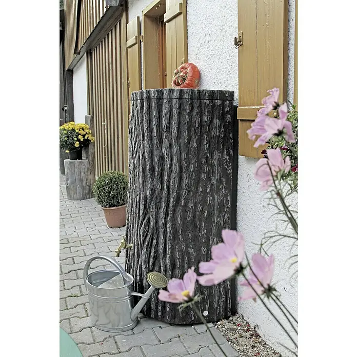 Arves Regenspeicher Evergreen Lite - 300 l, Dunkelbraun - Ein Regenfass in Baumstammoptik mit einem Kürbis darauf und einer Gießkanne davor, daneben rosa Blumen.
