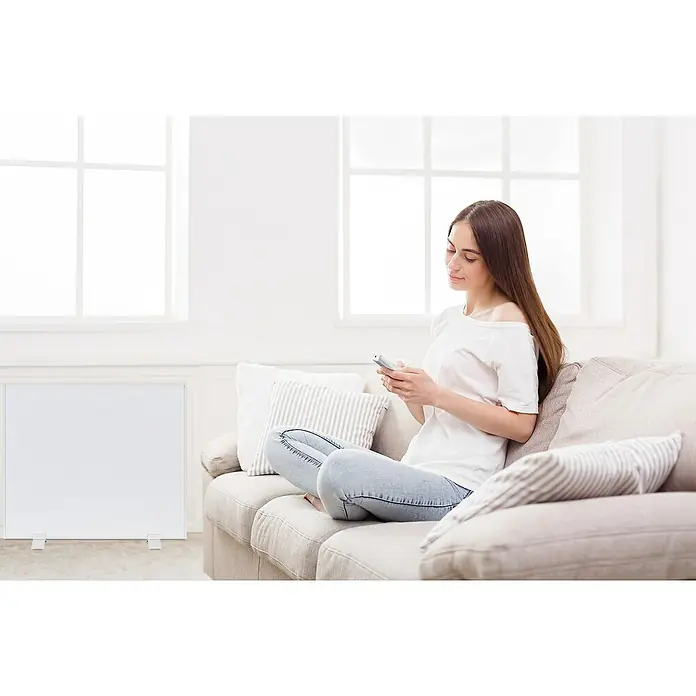 E2 elektro Infrarotheizkörper Complete Frame M - B x H: 600 x 50 cm, Weiß, 700 W - Eine junge Frau sitzt auf einem Sofa und benutzt ihr Handy. Im Vordergrund steht ein weißes Heizgerät.