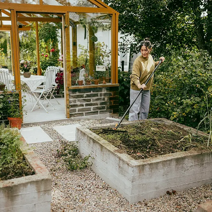 Fiskars OneClick Rasenrechen - L x B x H: 13 x 42 x 8,5 cm, 16 Stk. - Eine Frau harkt ein Hochbeet im Garten, während im Hintergrund ein Gewächshaus mit Tisch und Stühlen steht.