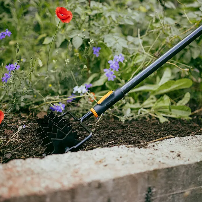 Fiskars OneClick Sternfräse - Arbeitsbreite: 21,2 cm, Stahl - Nahaufnahme eines Fiskars-Gartenwerkzeugs mit rotierenden Zinken, das in einem Blumenbeet verwendet wird.