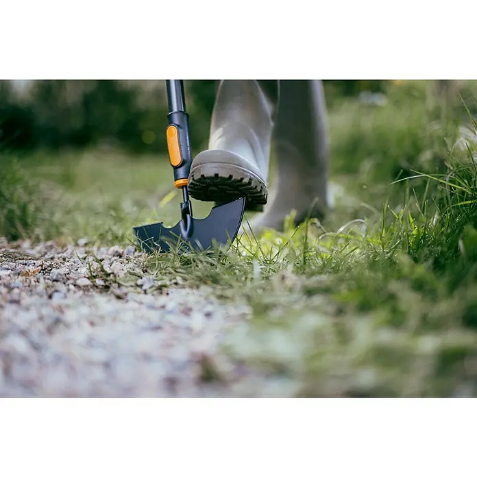 Fiskars OneClick Rasenkantenstecher - Arbeitsbreite: 19,7 cm, Stahl - Nahaufnahme eines Fußes in einem Gummistiefel, der auf einen Spaten mit orangefarbenem Griff in einem Garten tritt.