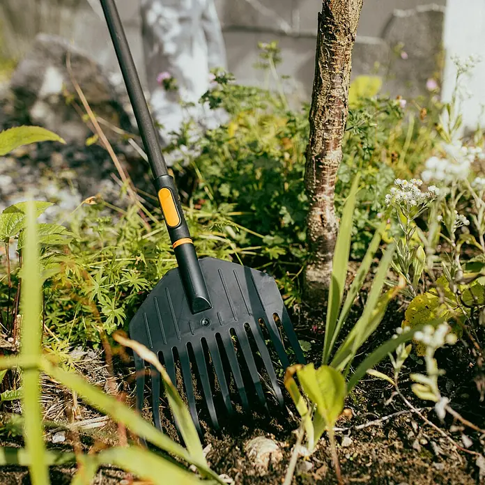 Fiskars OneClick Fächerbesen - Arbeitsbreite: 21,7 cm, Anzahl Zinken: 11 Stk. - Nahaufnahme eines schwarzen Fiskars-Laubfächers mit orangefarbenen Akzenten in einem Gartenbeet mit Pflanzen.